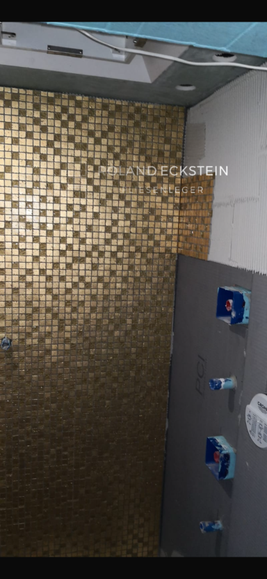 Dusche mit goldenem Fliesenmuster und blauen Wasserhähnen an der Wand.