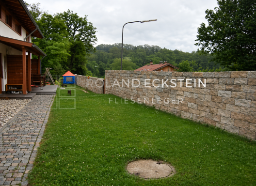 Grüner Rasen vor einer Steinmauer, links ein Holzgebäude und ein Zelt.
