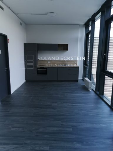 Leerer Raum mit Holzfußboden, großen Fenstern und moderner Kücheneinheit.