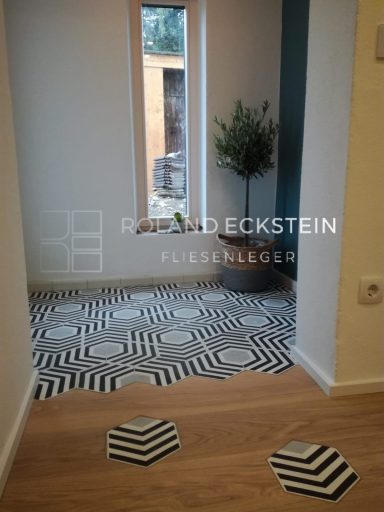Raum mit gemustertem Bodenbelag, Fenstern und einer Pflanzen im Hintergrund.