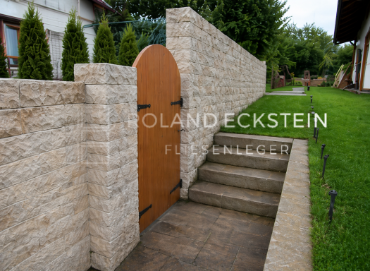 Holzzaun mit Rundbogen-Tür und Steinmauer, umgeben von Rasen und Bäumen.